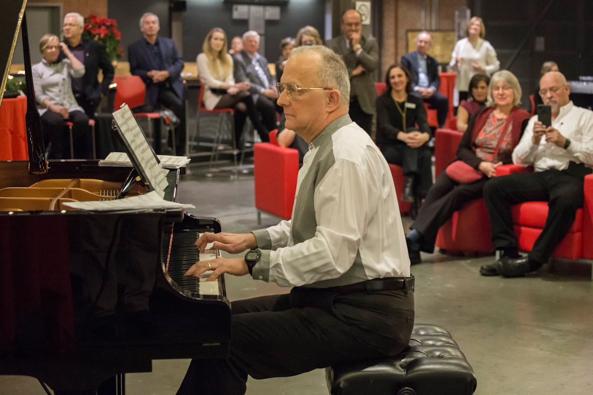 A grey haired Rafael Alcolado plays the piano as people look on.